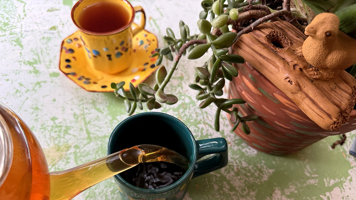 Pouring tea into a hand-painted teal Molly Mug beside a golden Octavia teacup, jade plant, and ceramic bird planter